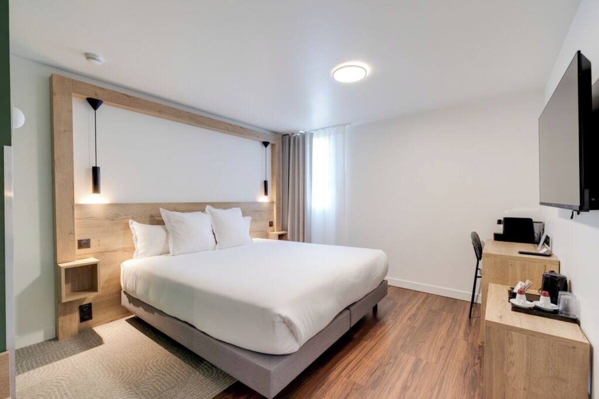 A modern hotel room with a large bed, wooden headboard, bedside tables, a desk, and a flat-screen TV.
