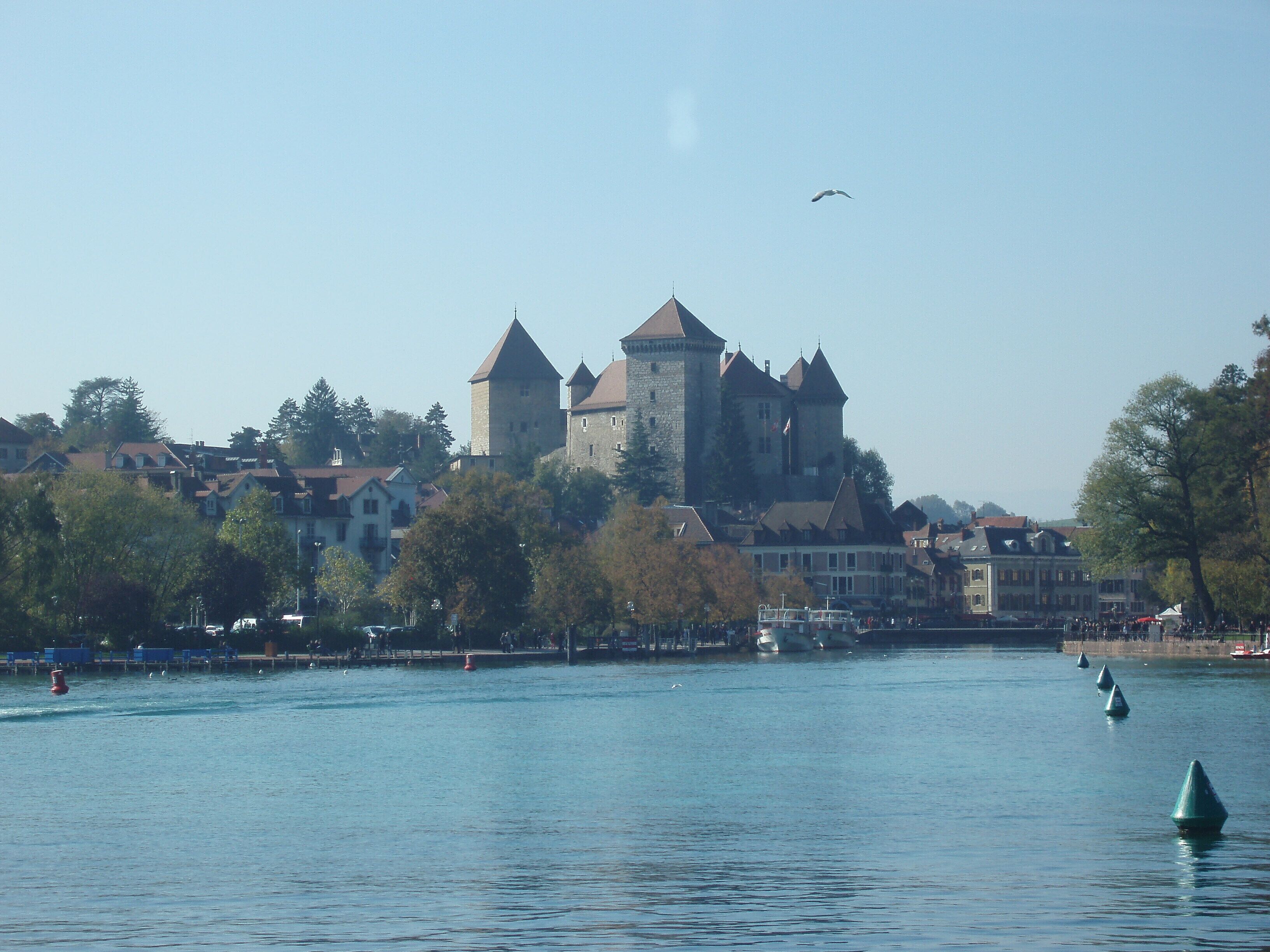 A historic castle with multiple towers, situated by a calm river with boats and buoys.