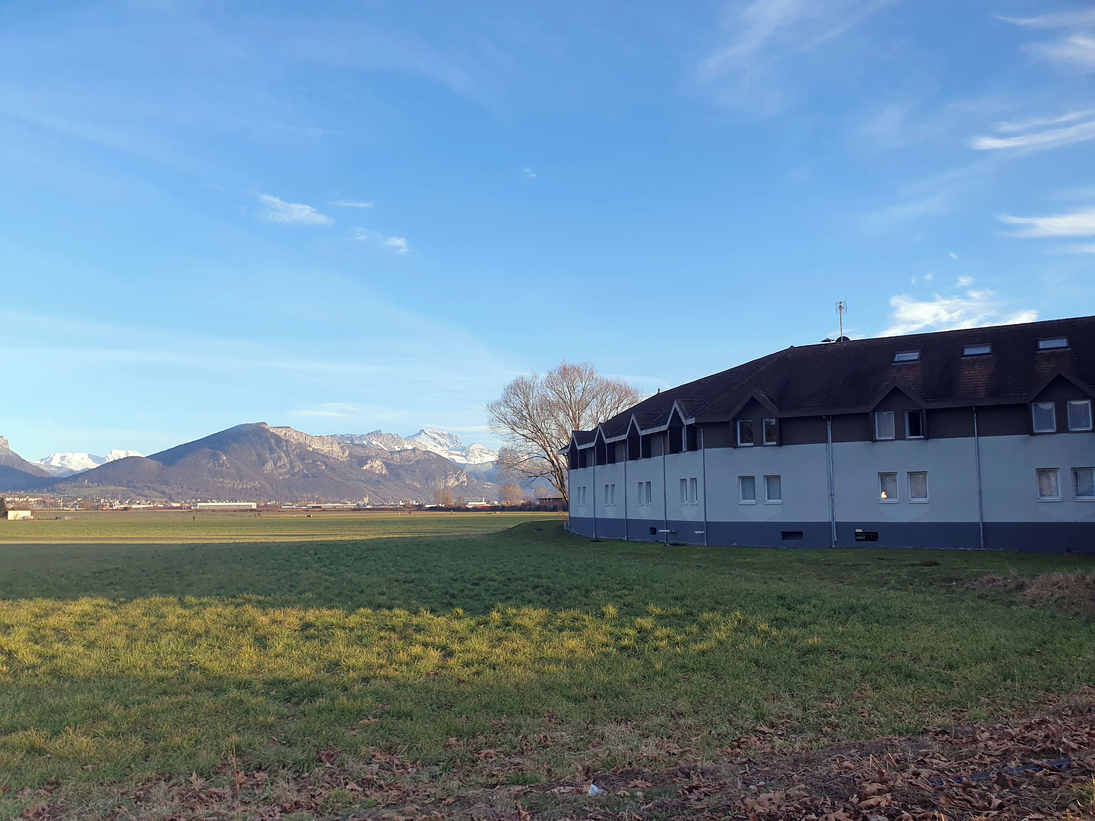 A large building with multiple windows and a flat roof, situated in a grassy field with mountains in the background.