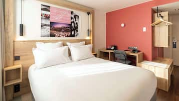 Quarto de hotel com uma cama grande, uma escrivaninha com cadeira, um cabideiro e uma obra de arte pendurada na parede.