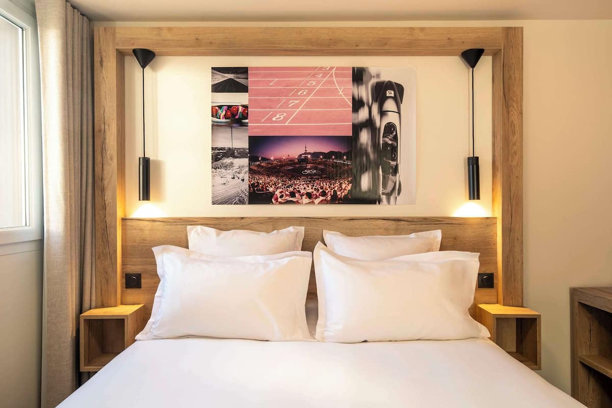 A neatly made bed with white linens and pillows, flanked by two bedside tables with lamps. Above the bed, there is a wall-mounted artwork featuring various photographs and a chalkboard with numbers.
