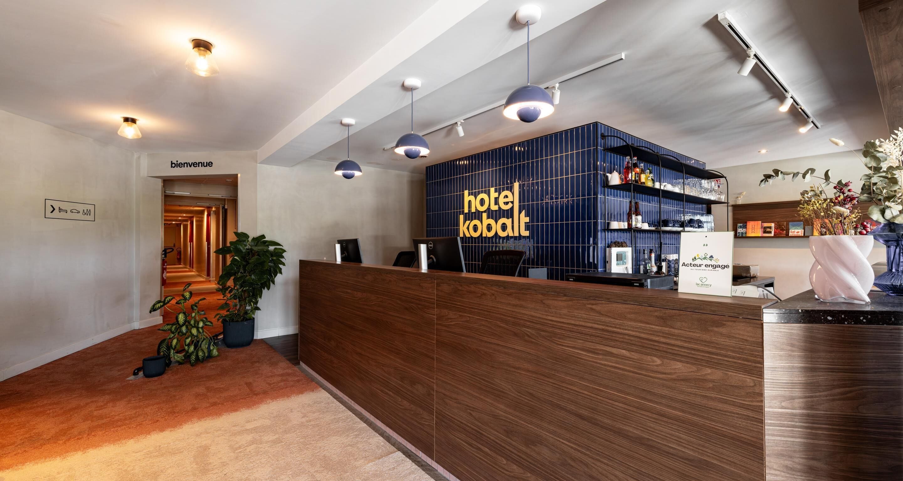 Un mostrador de recepción de hotel con encimera de madera, una pared azul con el nombre del hotel, una zona de recepción con plantas en macetas y un cartel que indica la entrada.