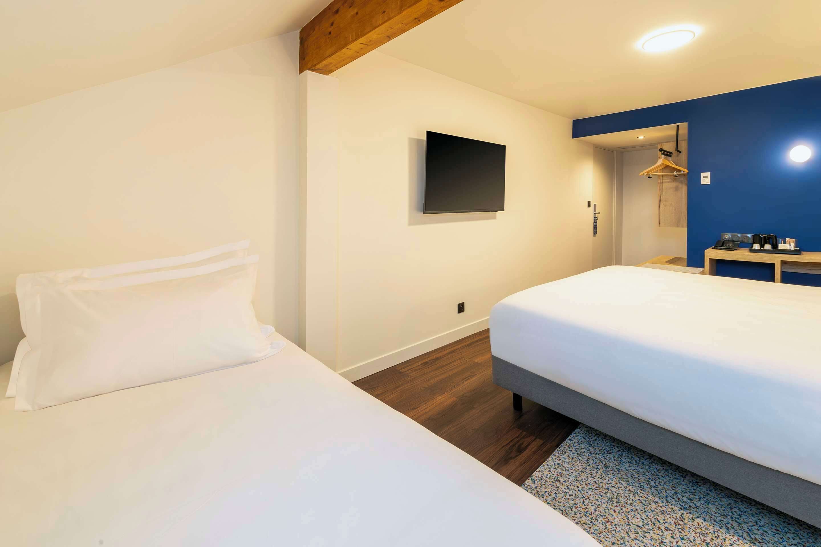 Habitación de hotel con dos camas, un televisor de pantalla plana montado en la pared, una viga de techo de madera, y un escritorio pequeño con un espejo y una cafetera.