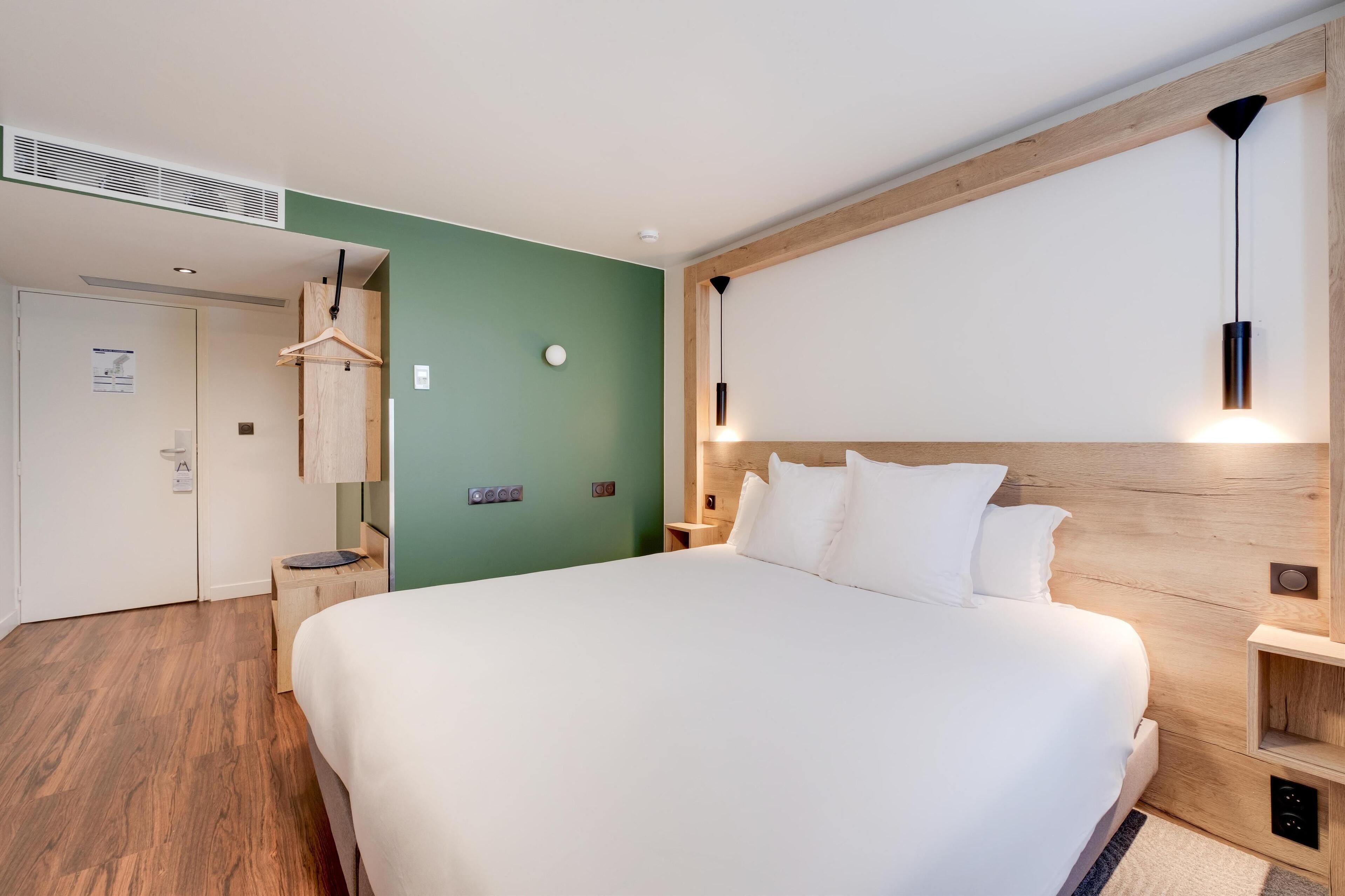 Une chambre d’hôtel moderne avec un grand lit, une tête de lit en bois, un mur d’accent vert, un luminaire suspendu et une petite table de chevet.
