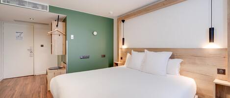 Una habitación de hotel moderna con una cama grande, un cabecero de madera, una pared verde de acento, un colgador de luz y una mesita de noche pequeña.