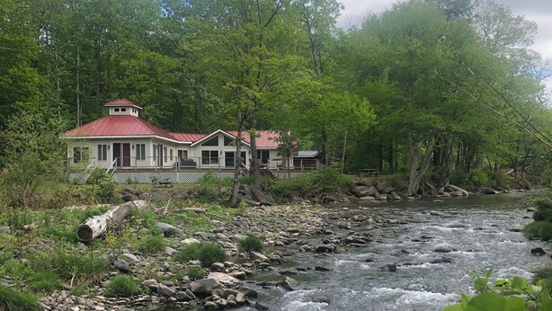 A riverside cabin with a red roof, surrounded by dense forest and a flowing river.