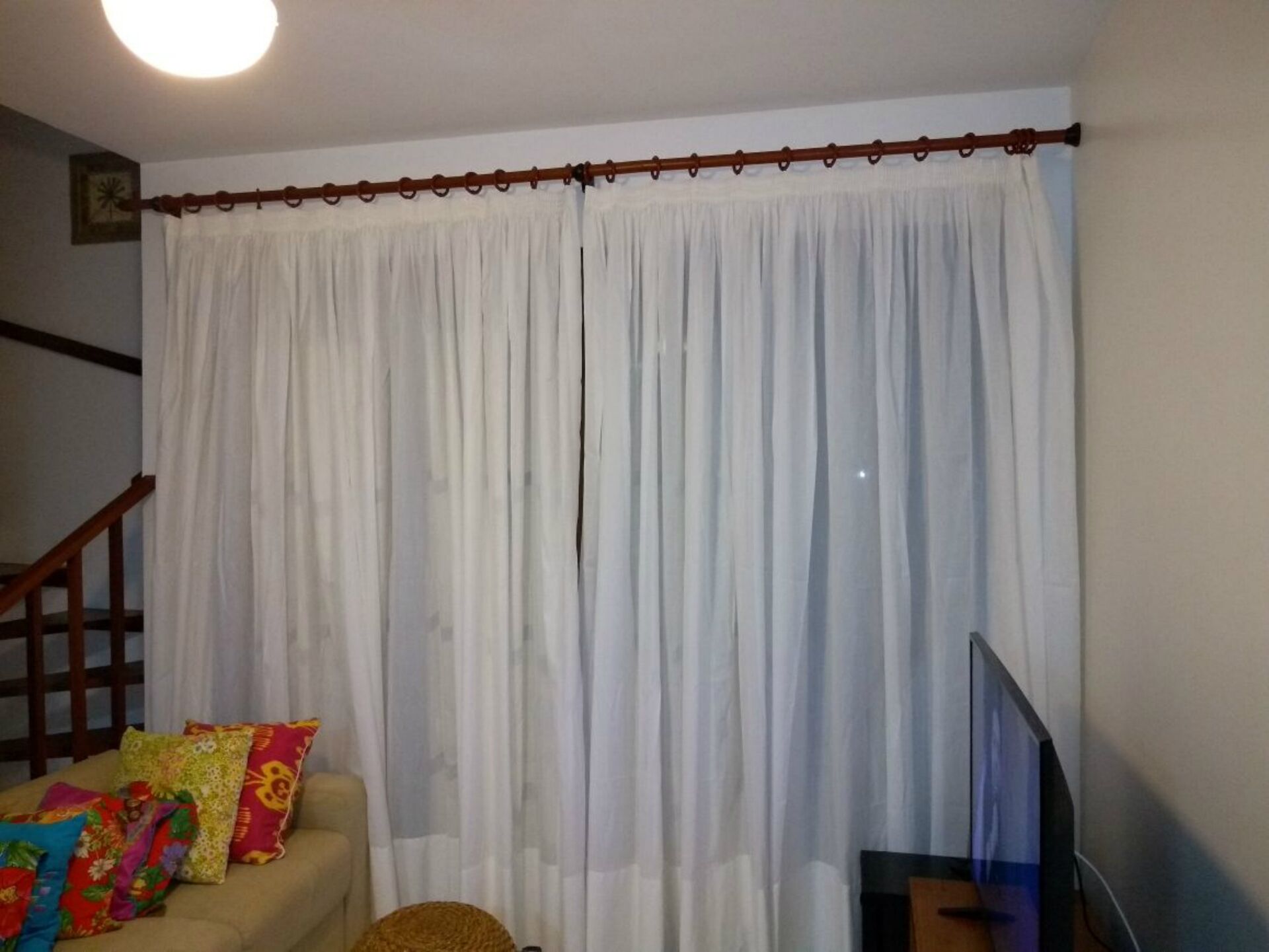 Ein Wohnzimmer mit einer Couch, farbenfrohen Kissen, einem Fernseher und einem großen Fenster mit weißen Vorhängen.