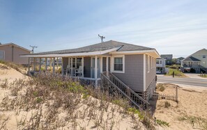 A single-story house with a porch, a staircase leading to the entrance, and a sandy beach in the background. - Wayfarer's Rest - Kitty Hawk MP 3.5, Oceanfront, Ocean View, Year Round Flex Stays (Kitty Hawk)