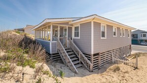 Una casa frente a la playa con un porche cubierto, una escalera que lleva a la entrada y una playa de arena al fondo.