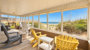 A covered deck with wooden flooring, outdoor seating including Adirondack chairs and a table, and large windows offering a view of the beach and ocean.