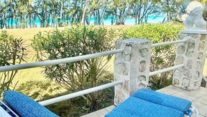 Una silla de descanso con cojín azul en un patio empedrado, con vistas a una playa con palmeras y cielo despejado.
