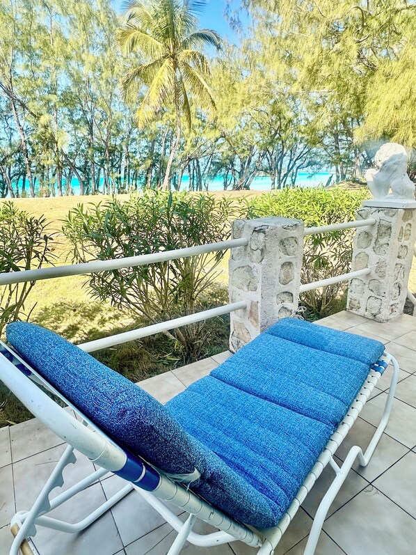 A lounge chair with a blue cushion on a tiled patio, overlooking a beach with palm trees and a clear sky.