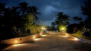 A paved road flanked by stone walls and palm trees, leading to a building with illuminated entrance. - Stunning Luxury Villa Directly On French Leave Beach (Governors Harbour)