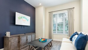 A living room with a flat-screen TV mounted on the wall, a wooden cabinet, a dining table, and a beige sofa with blue cushions.