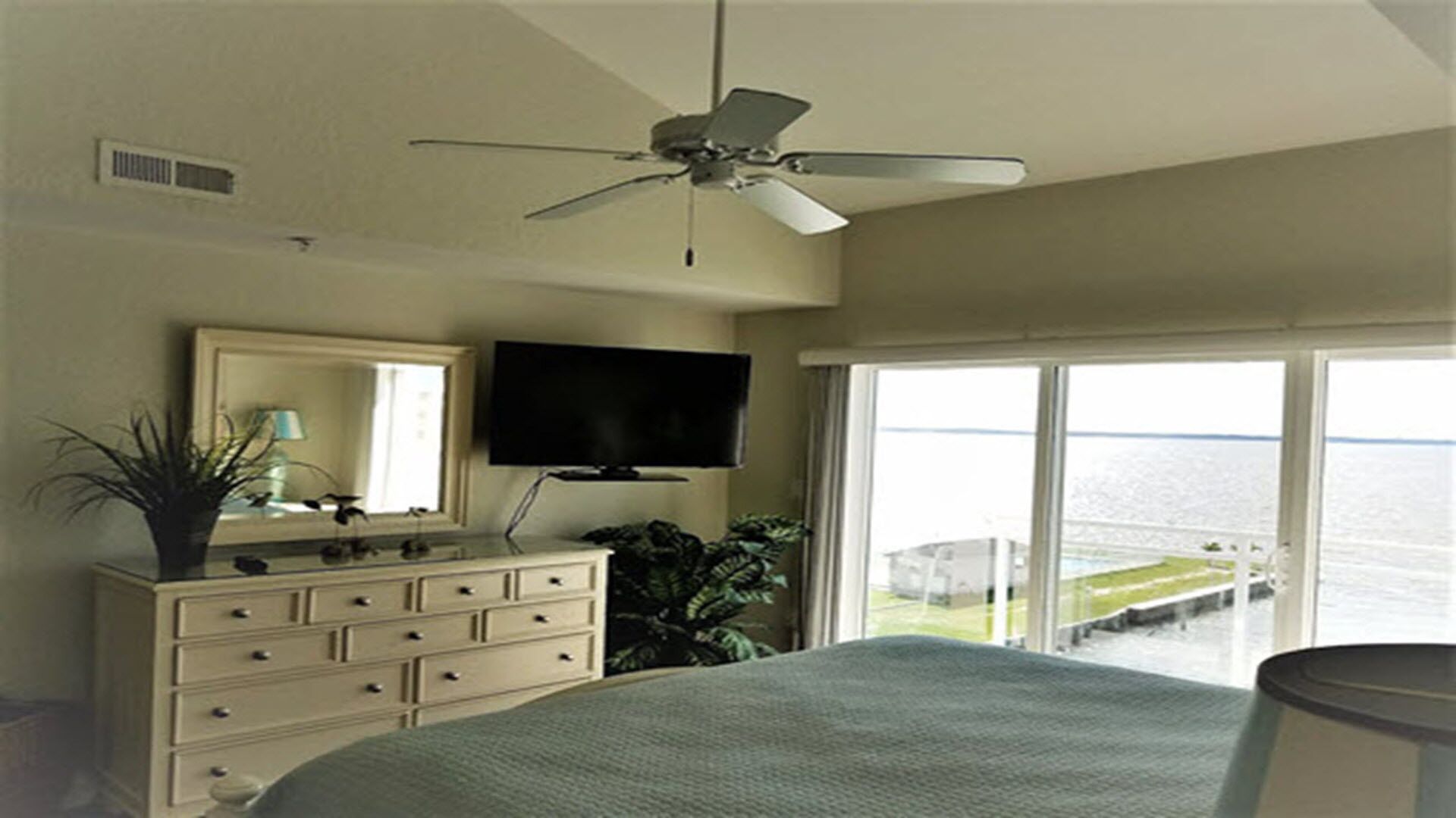 A bedroom with a bed, dresser, TV, and a balcony with a view of the water.