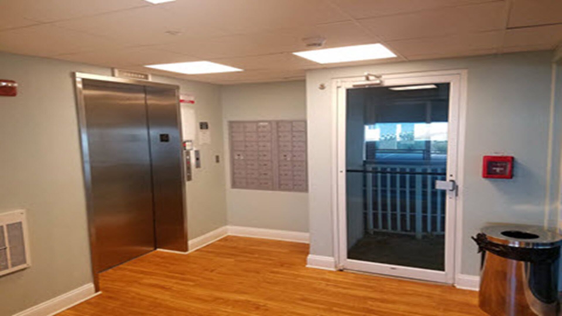 An elevator, a door with a glass panel, a trash can, and a bulletin board in a hallway.