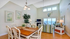 A dining area with a wooden table and white chairs, a large plant, and a view of the ocean through the windows. - 402SunsetBeach,PenthouseDir.WaterFrt,Pvt.Beach,Pool,Elev,1Blk Seacrets3BDRM/3FBH (Ocean City)