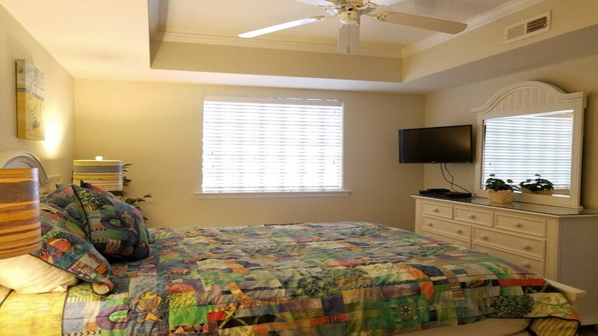 Une chambre à coucher avec un lit, un ventilateur de plafond, une télévision et une fenêtre avec des stores.