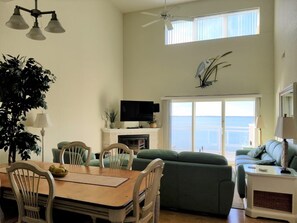 Un salon comprenant une table à manger, des chaises, un canapé vert, un téléviseur et une vue sur la mer grâce à de grandes fenêtres.
