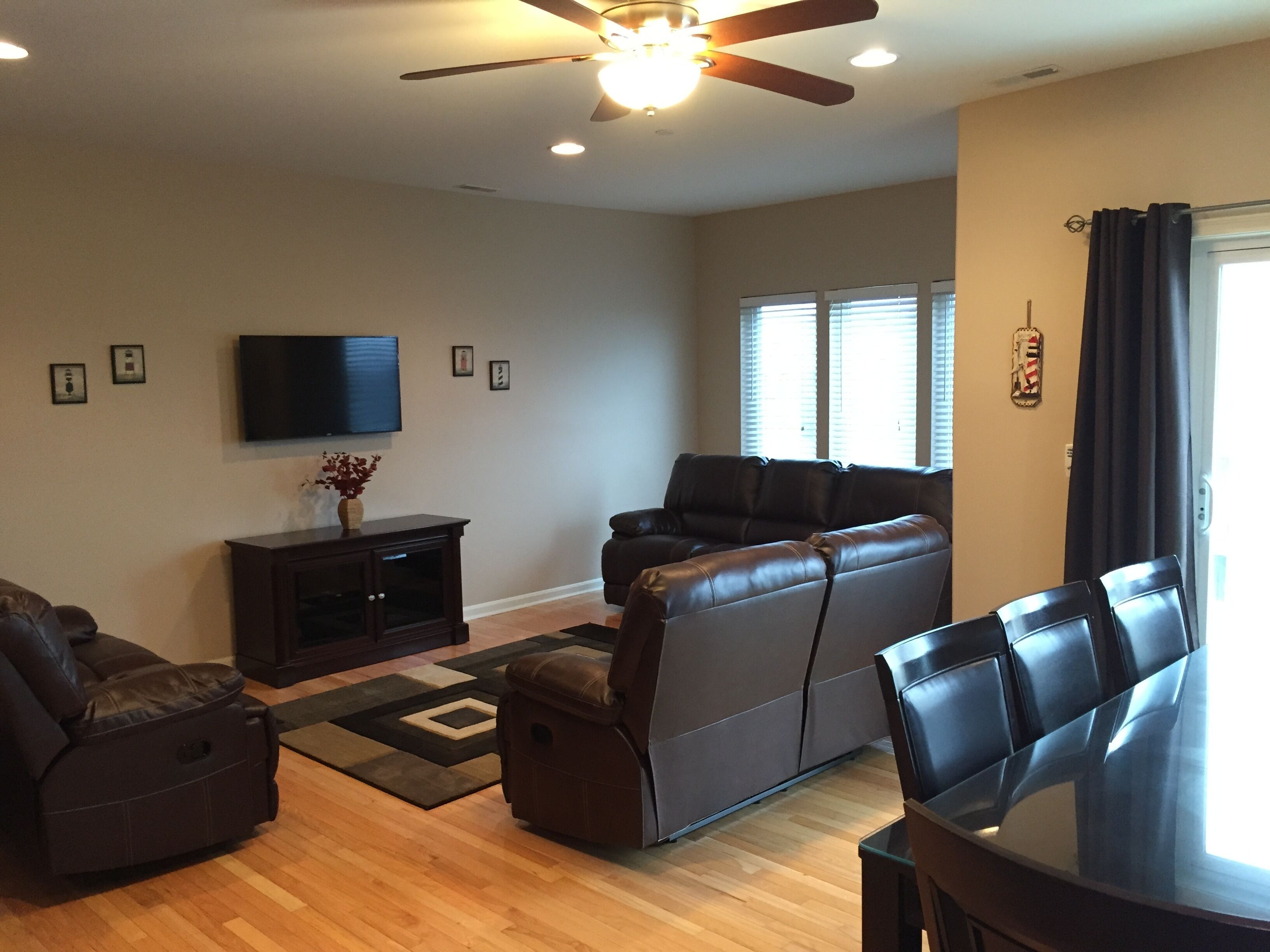 A living room with a dark brown leather sofa, a brown leather chair, a glass-top dining table, and a ceiling fan.