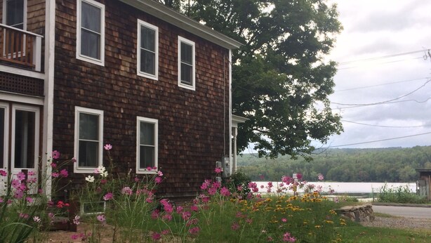 Une maison en bois à deux étages, dotée d’un balcon, entourée d’un jardin où poussent différentes sortes de fleurs et bordée d’un mur de pierre.