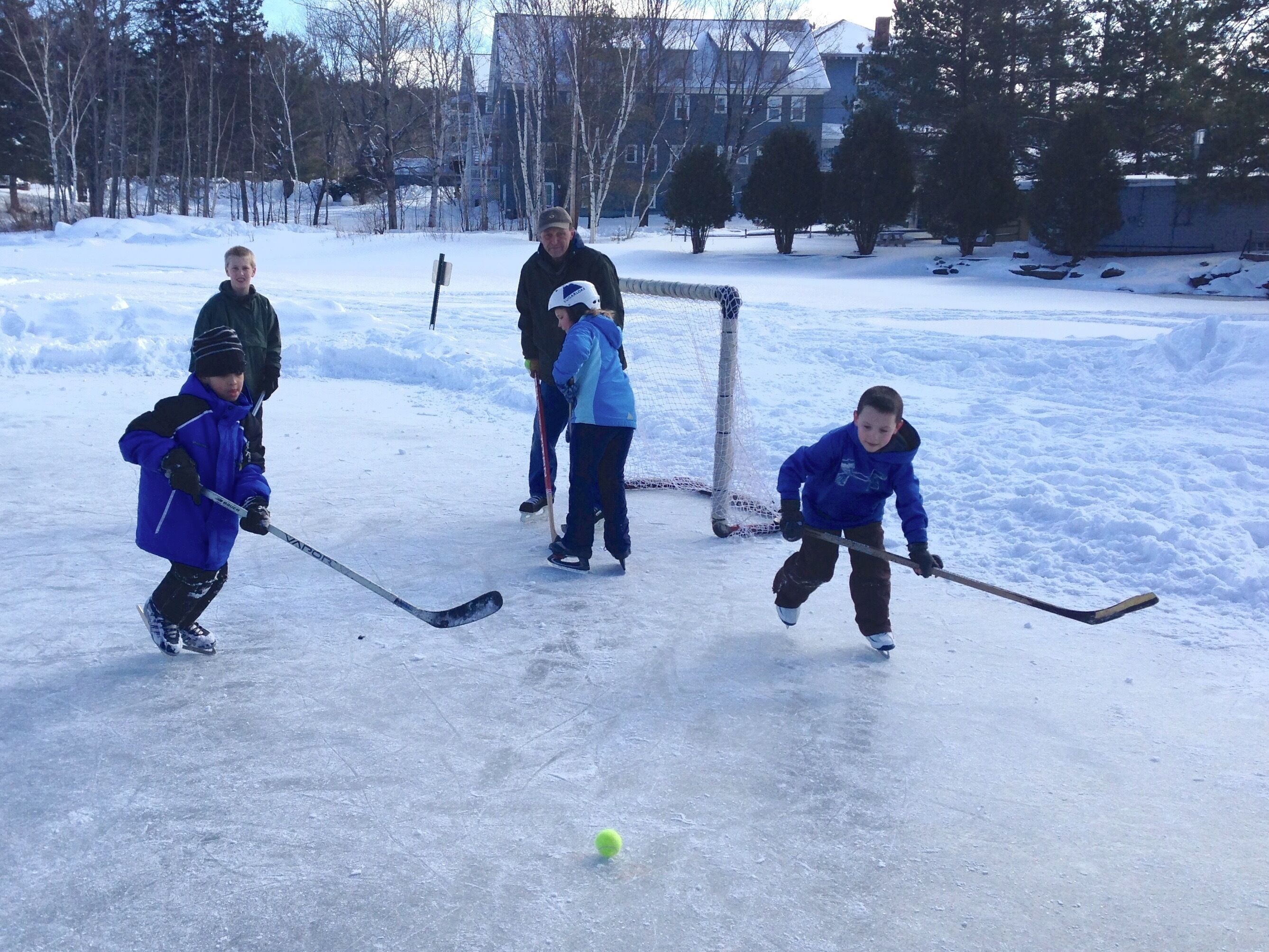 A group of children playing hockey on an outdoor ice rink surrounded by trees and buildings.