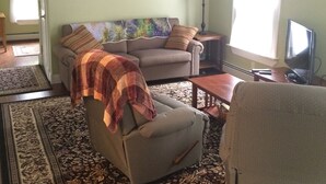 Un salon avec un canapé, un fauteuil et un téléviseur. Il y a un ventilateur de plafond et une fenêtre avec des rideaux.