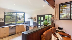 A modern kitchen with wooden cabinets, a dishwasher, and a large window. There is a dining area with a brown leather couch and a vase on the table. - Tranquil Oasis - Pet friendly (BALNARRING BEACH)