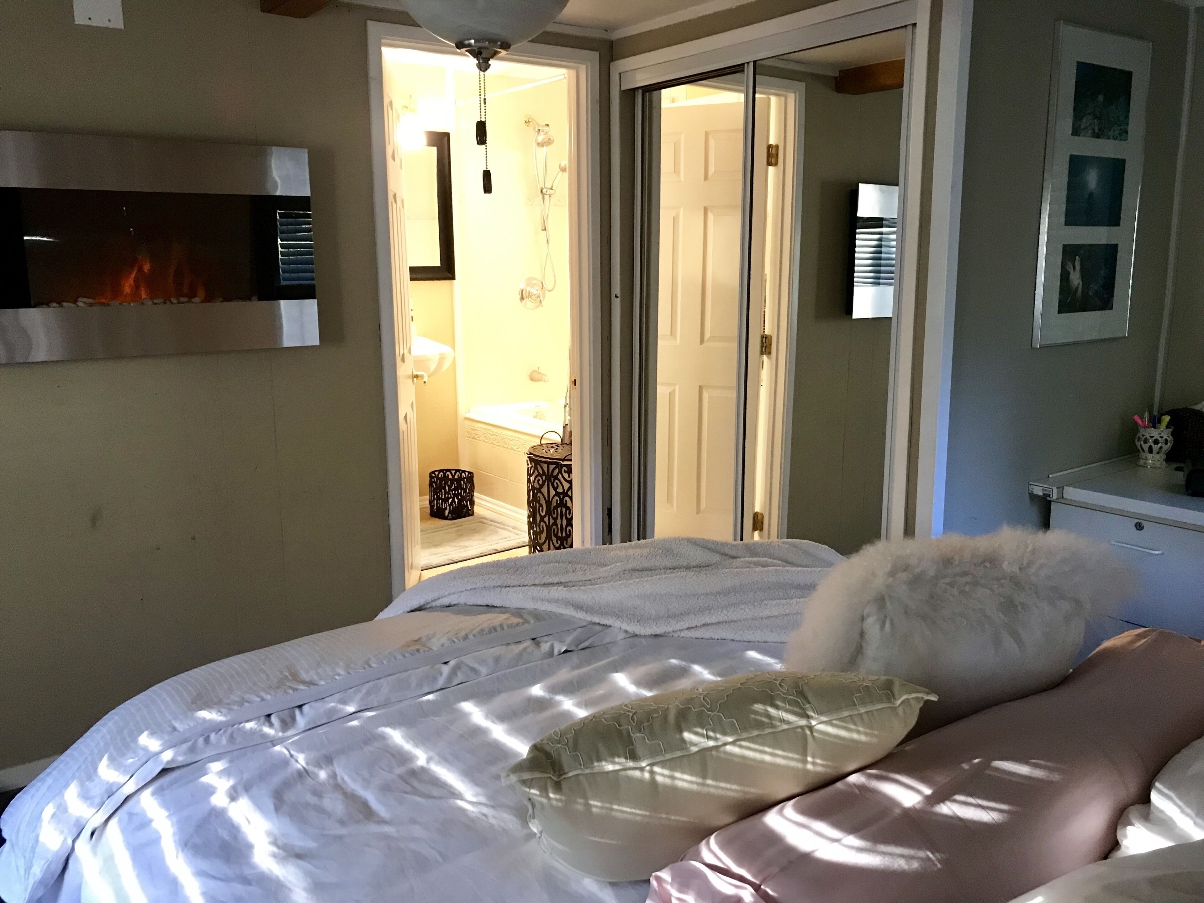 A bedroom with a bed, a fireplace, a bathroom visible through an open door, and a desk with a lamp.