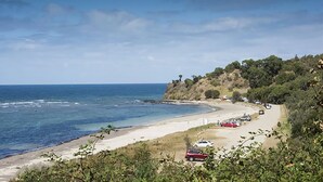 Een kustweg met geparkeerde auto's, een strand en een klif begroeid met begroeiing.