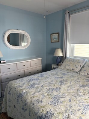 Un dormitorio con cama, cómoda, lámpara, espejo y ventana con cortinas.