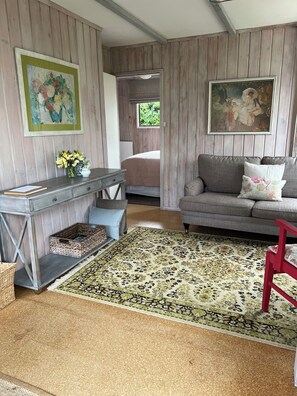 A room with a wooden paneled wall, a grey sofa, a red chair, a wooden console table with a vase of flowers, and a patterned rug. - Quiet, peaceful country cottage (Pukekawa)