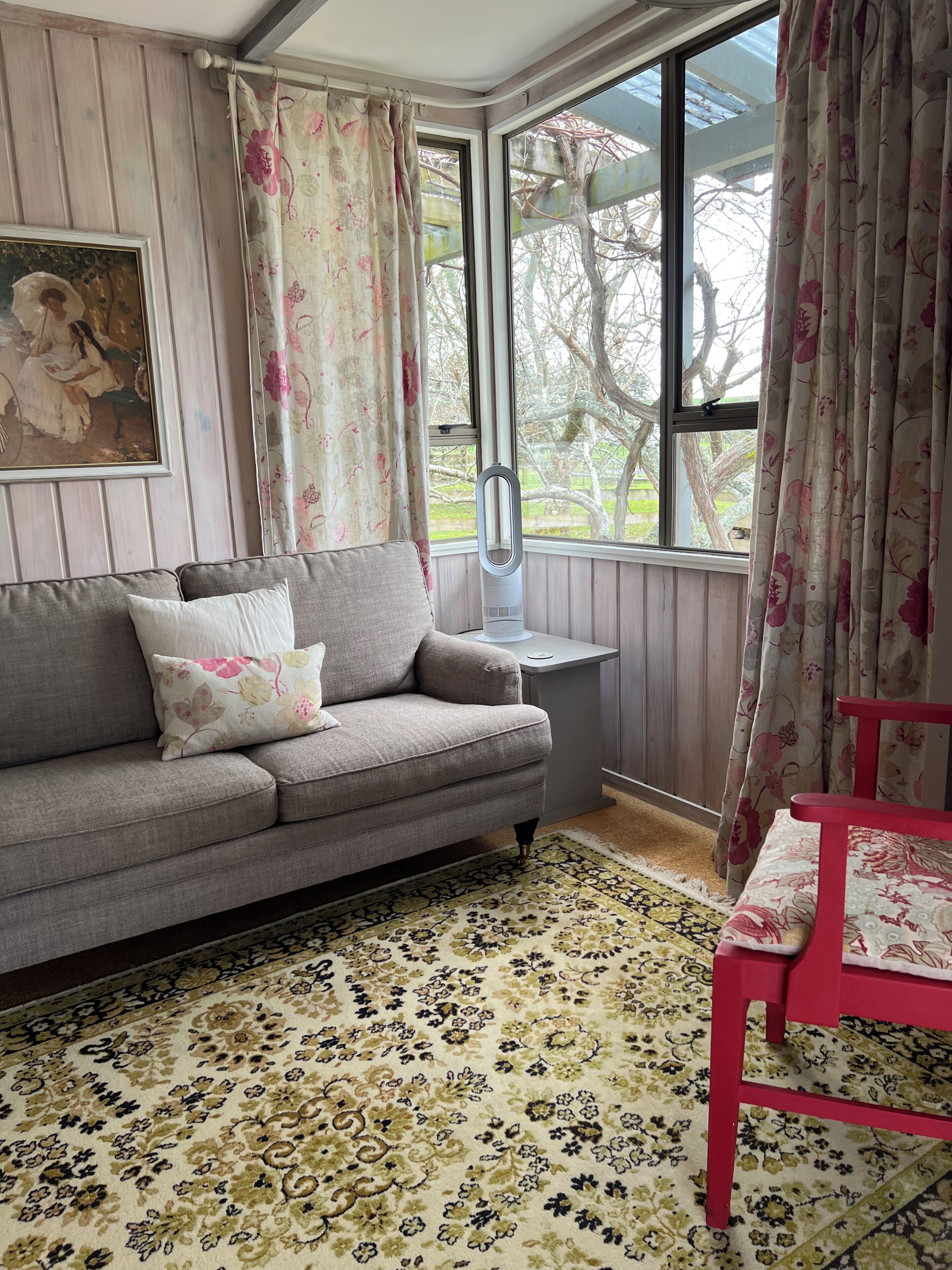 Um quarto com sofá cinzento, uma cadeira vermelha, um tapete estampado, um quadro na parede e uma janela com cortinas.