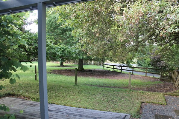A covered outdoor area with a wooden deck, surrounded by trees and a fenced path. - Quiet, peaceful country cottage (Pukekawa)