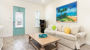 Ein Wohnzimmer mit einer türkisblauen Tür, einem beigen Sofa mit Kissen, einem Holztisch und einem Bild von einem tropischen Strand.