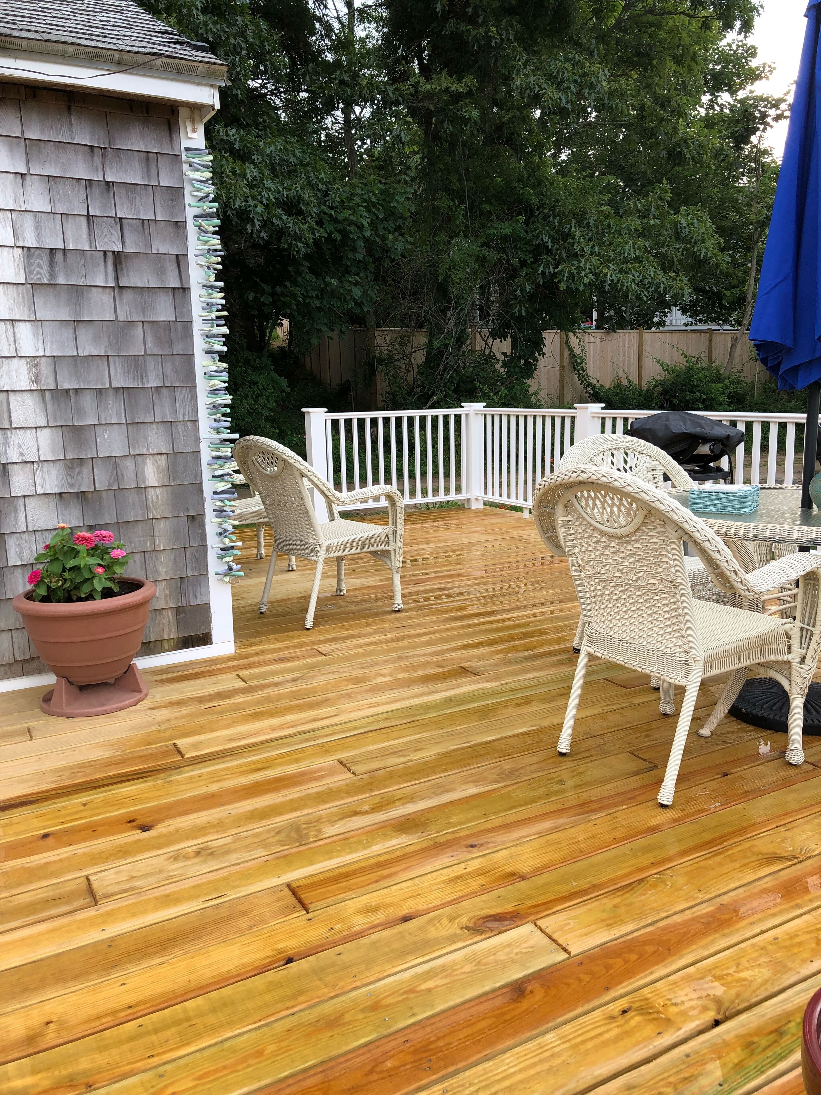 A wooden deck with white wicker chairs and a table, surrounded by a white fence and trees.