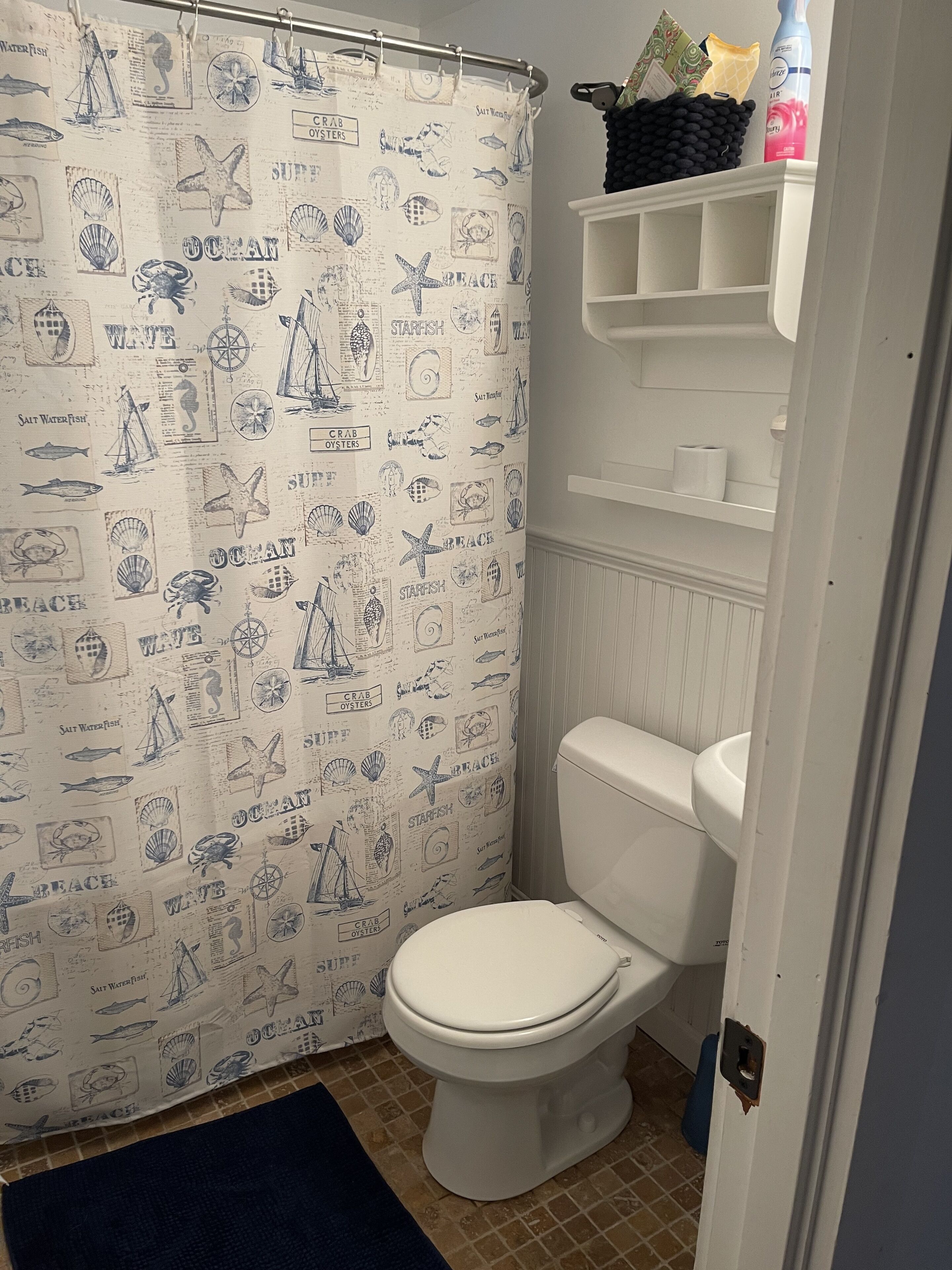 Une salle de bain avec un rideau de douche à motifs nautiques, des toilettes blanches et une étagère contenant des articles de toilette.