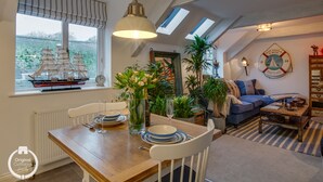 Un comedor con una mesa de madera, sillas blancas y un modelo de barco en el alféizar de la ventana. Hay plantas en macetas y un sofá azul en la sala de estar.