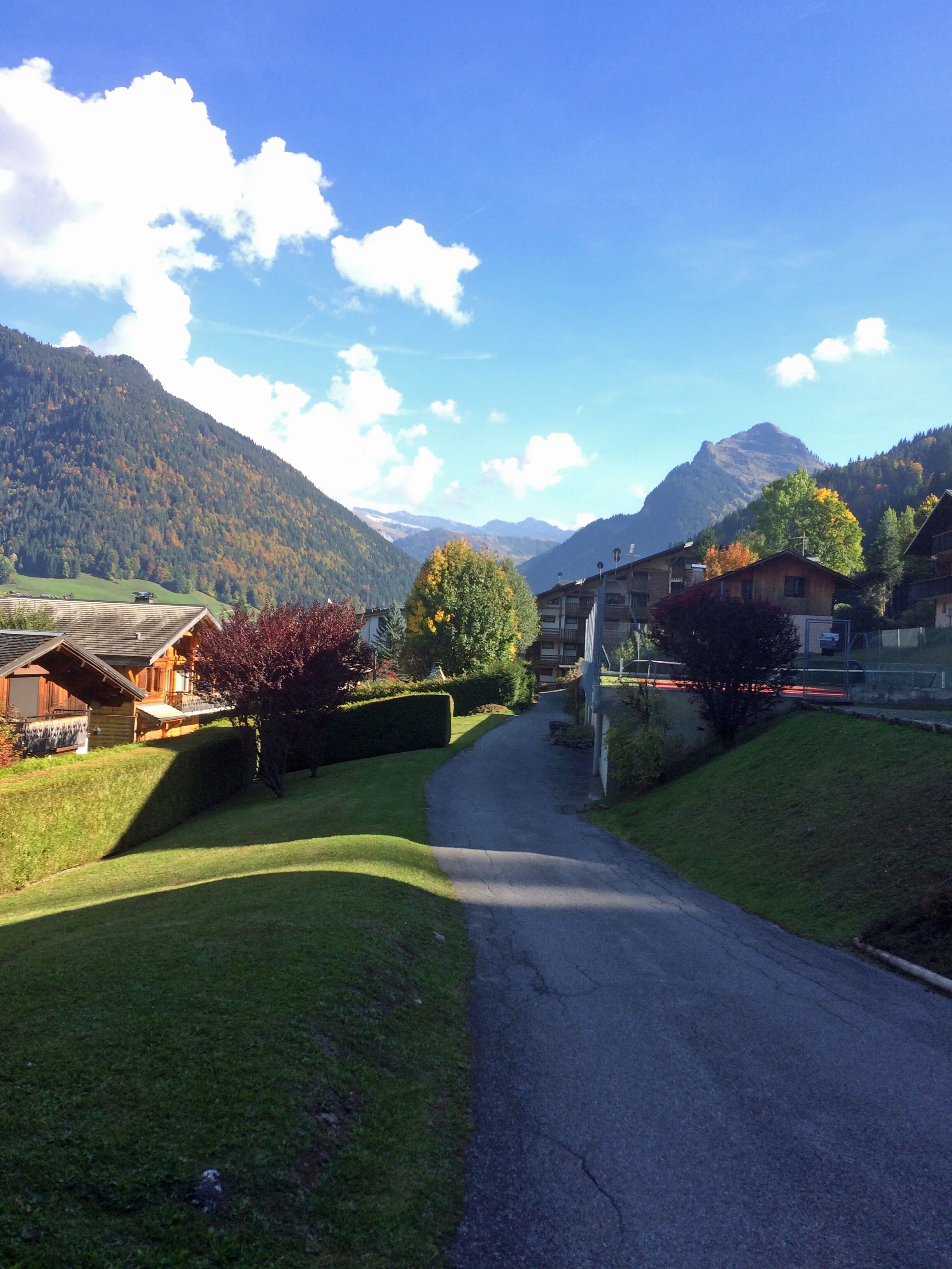 Eine Landstraße führt zu einem Dorf mit traditionellen Holzhäusern, umgeben von Bergen und Herbstlaub.