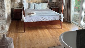 Een slaapkamer met een hemelbed, stenen muren, houten vloer, een groot raam en een bad.