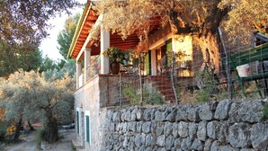 A stone-walled building with a wooden balcony, surrounded by trees and a stone pathway.