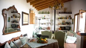 A cozy living room with a wooden dining table, chairs, and a chandelier. There are shelves with various items and a large mirror on the wall.