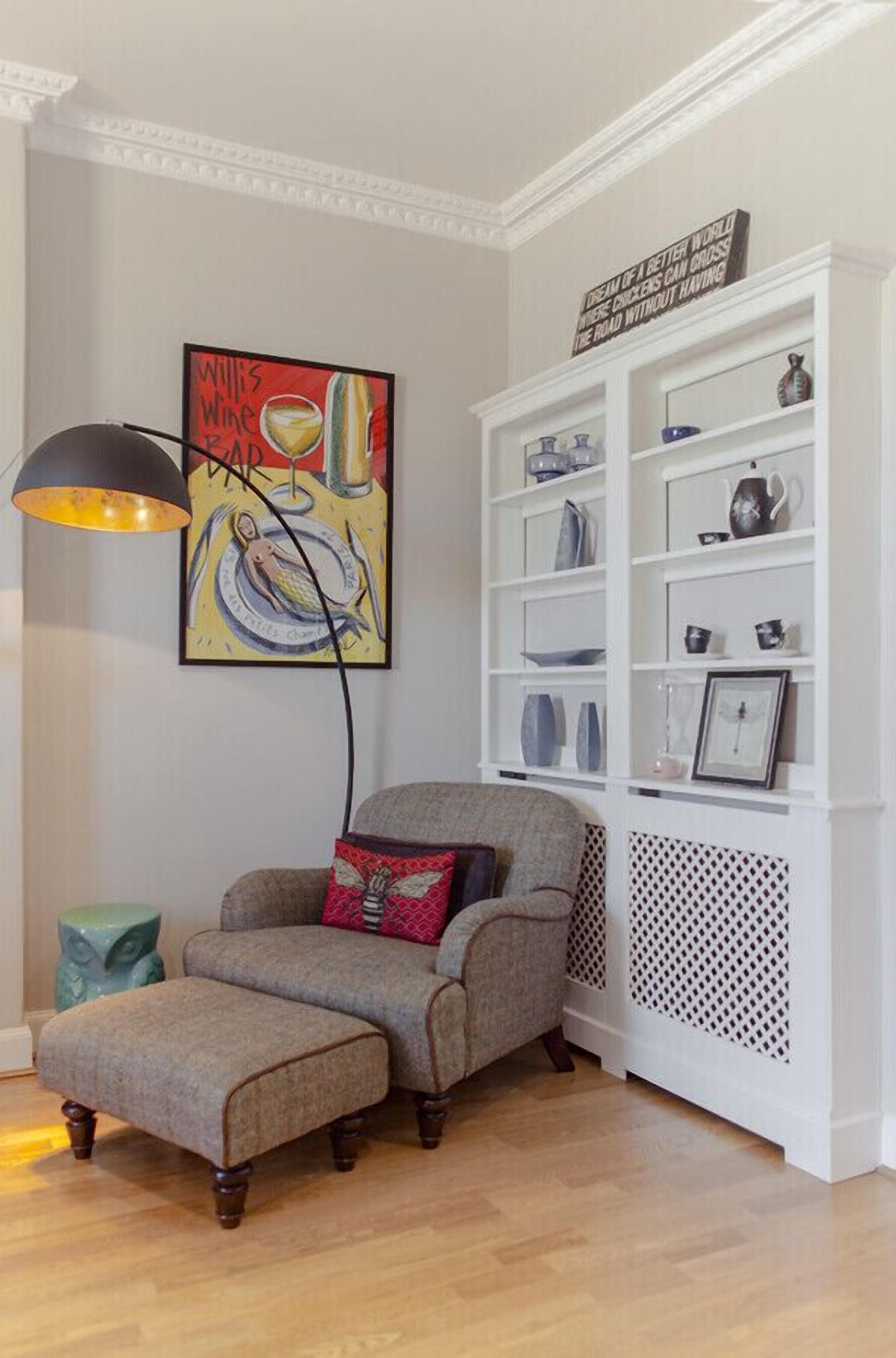 Une pièce comprenant un fauteuil gris, un coussin rouge, une étagère blanche contenant divers objets et une lampe fixée au mur.