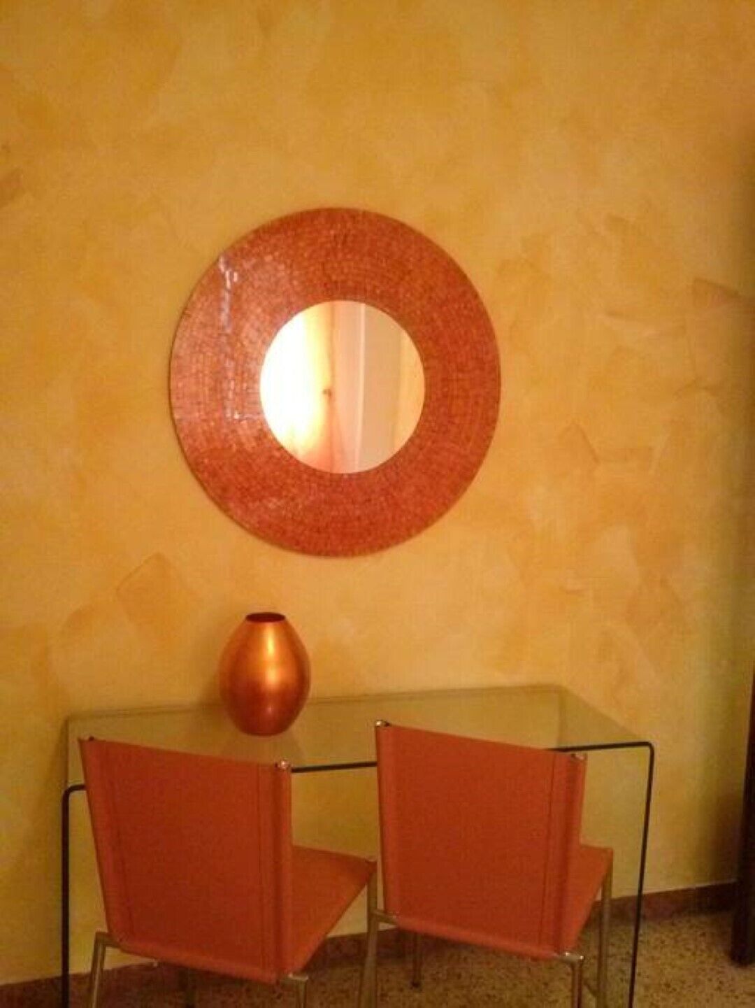 Una habitación con un espejo redondo en la pared, dos sillas rojas y una pequeña mesa con un florero.
