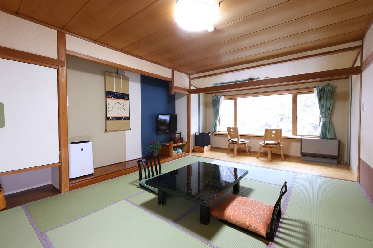 A traditional Japanese-style room with tatami flooring, a low table with cushions, a television, and a window with curtains.
