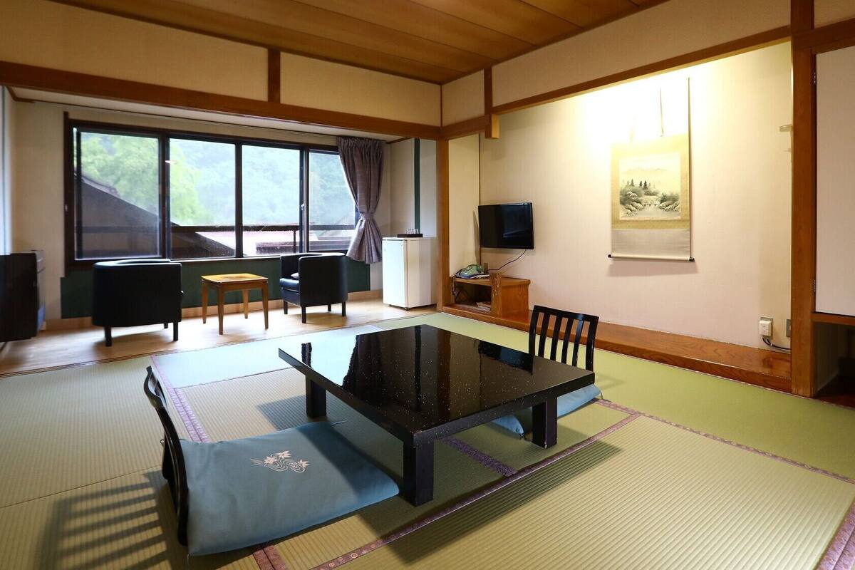 A traditional Japanese room with tatami flooring, a low table, chairs, a television, and a window with a view.