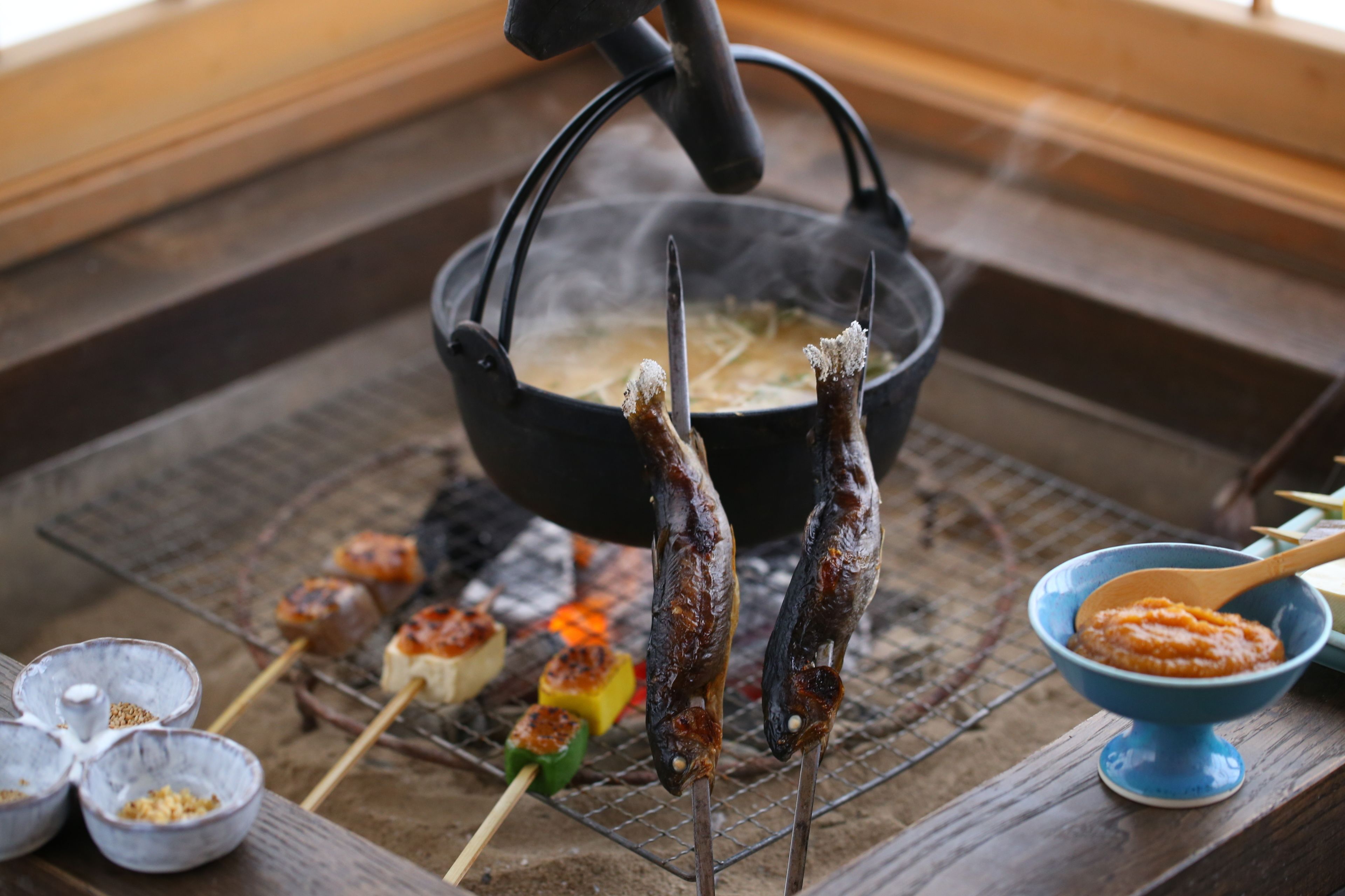 A grill with skewers of meat and vegetables, a pot of boiling water, and small bowls of condiments.