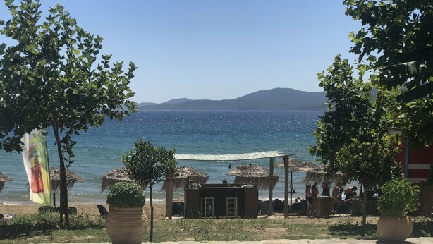 Vista de platja amb cel serè, mar tranquil·la i muntanyes llunyanes. Hi ha sombrerets de palla i plantes en testos.
