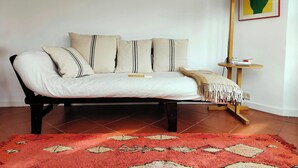 A bed with a white sheet, multiple pillows, a red patterned rug, a floor lamp, and a small table with books.
