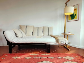 Un lit avec un drap blanc, plusieurs oreillers, un tapis rouge à motifs, une lampe sur pied et une petite table avec des livres.